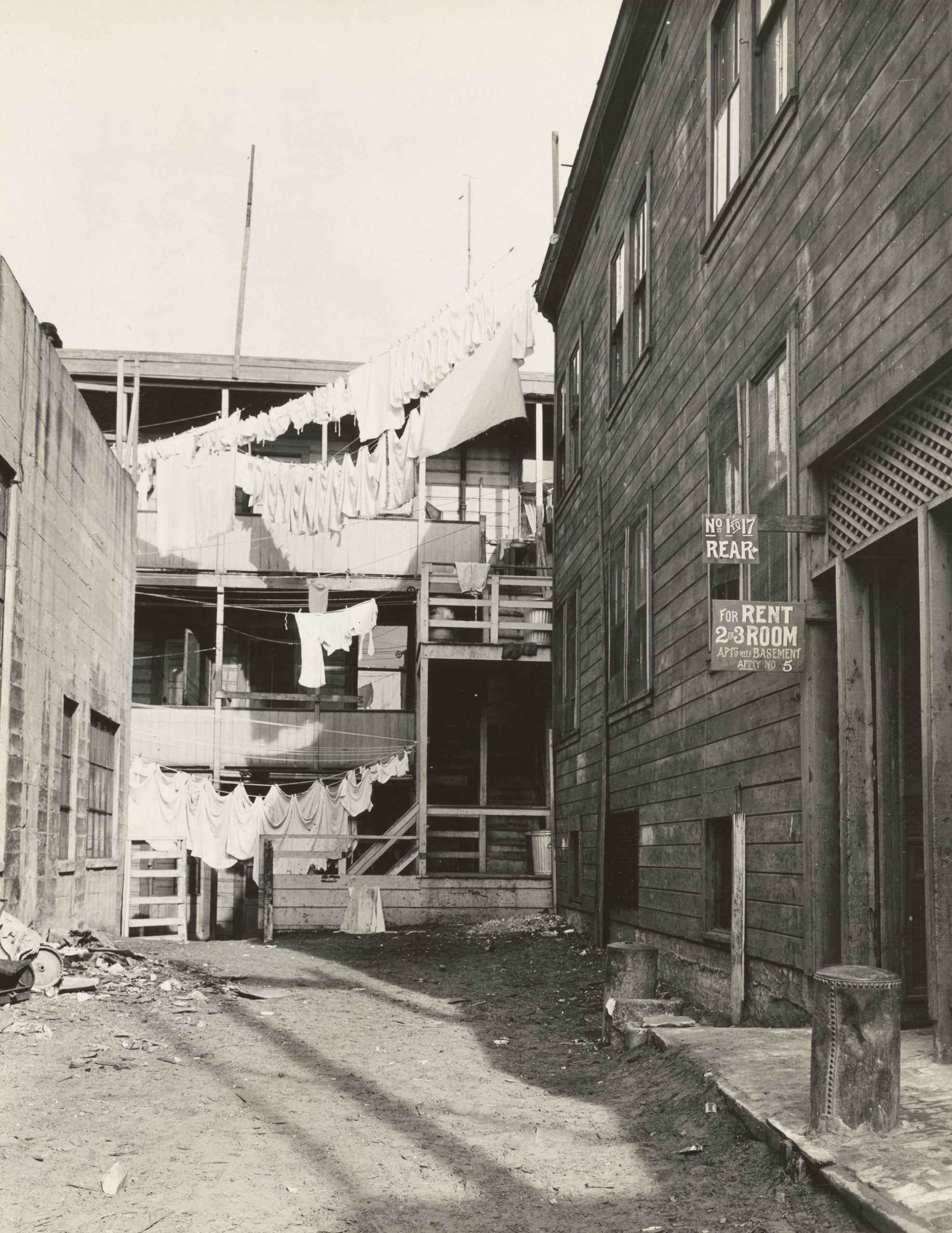 Washing strung on lines across a tenement rear yard, beside a sign reading For Rent 2-3 Room